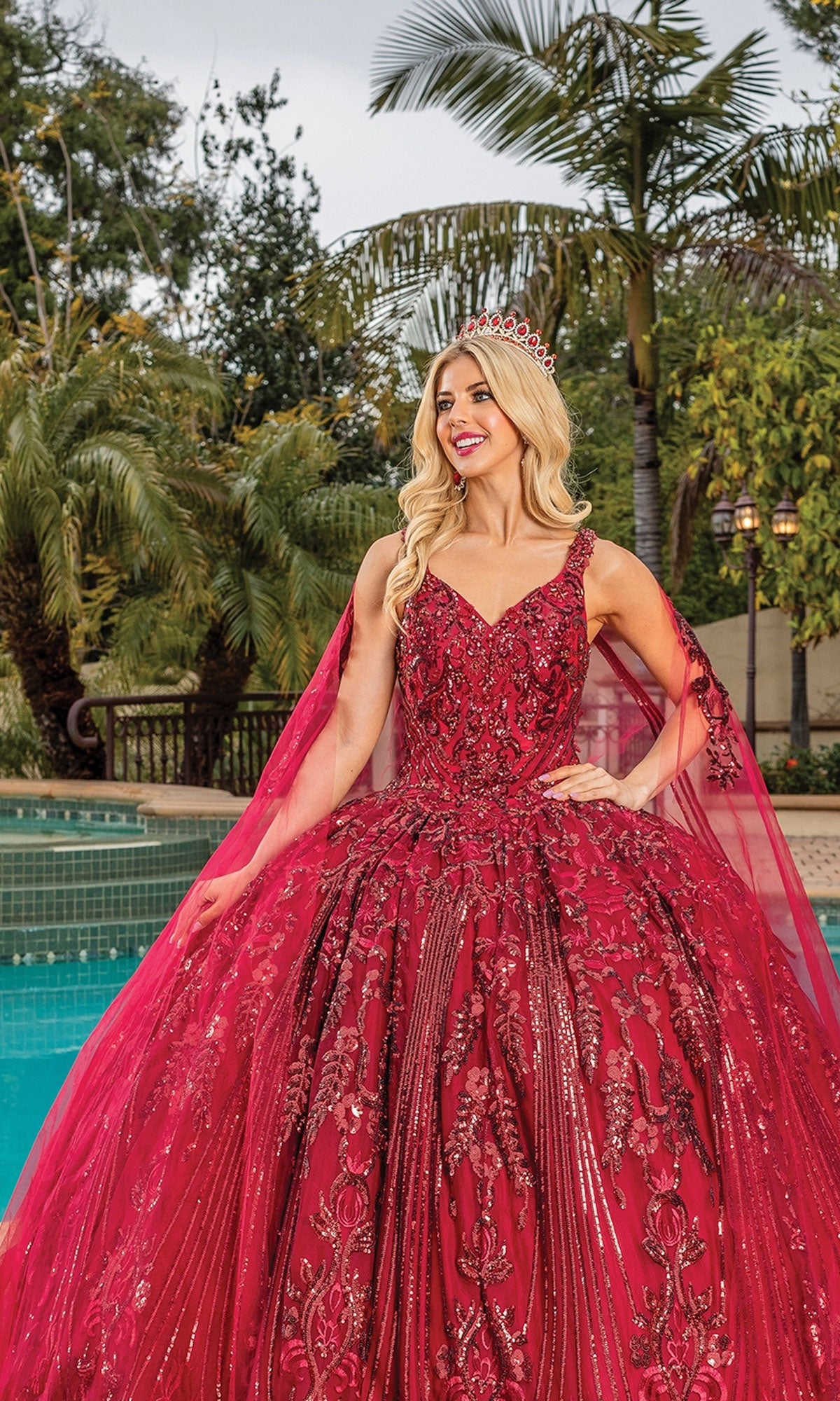 A woman wearing the Quinceanera Dress 1649 By Dancing Queen, featuring a red ball gown and sweetheart neckline with a tiara, stands before a swimming pool with palm trees in the background.